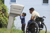 Spanischer Mann mit Querschnittslähmung im Rollstuhl, sein Sohn schaut in einen Briefkasten — Stockfoto