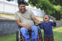 Homme hispanique avec blessure à la moelle épinière en fauteuil roulant avec son fils riant dans la pelouse — Photo de stock