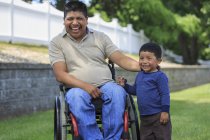 Uomo ispanico con lesione del midollo spinale in sedia a rotelle con suo figlio che ride nel prato — Foto stock