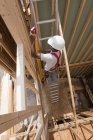 Hispanic carpenter carrying a level up ladder to second floor at a house under construction — Stock Photo