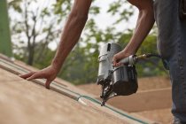 Hispanic carpenter using a nail gun on the roof panel of a house under construction — Stock Photo