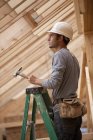 Carpinteiro hispânico em pé em uma escada com um martelo em uma casa em construção — Fotografia de Stock