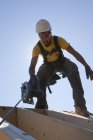 Carpintero hispano usando una sierra circular en las vigas del techo de una casa en construcción - foto de stock
