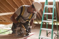 Hispanic carpenter using a circular saw on roof panel at a house under construction — Stock Photo