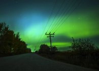 Northern Lights brillando verde en el cielo estrellado sobre una carretera rural y líneas de transmisión, Condado de Sturgeon; Alberta, Canadá - foto de stock