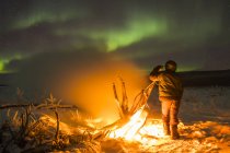 Оставаясь в тепле у костра на реке Дельта, наблюдая за северным сиянием в холодную ночь; Аляска, Соединенные Штаты Америки — стоковое фото