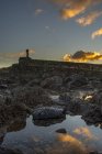 Un seul moai lointain est représenté contre le ciel éclairé par le lever du soleil, qui se reflète dans une piscine d'eau au premier plan ; Île de Pâques, Chili — Photo de stock