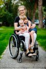 Una madre parapléjica llevando a sus hijas en su regazo mientras usa una silla de ruedas al aire libre en una cálida tarde de verano y posando para la cámara: Edmonton, Alberta, Canadá - foto de stock