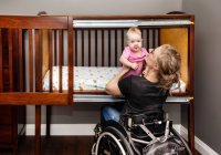 Eine querschnittsgelähmte Mutter hebt ein Baby aus einem maßgeschneiderten, seitlich öffnenden Gitterbett, das es ihr ermöglicht, ihr Baby für ein Nickerchen aus ihrer Position im Rollstuhl abzulegen: Edmonton, Alberta, Kanada — Stockfoto
