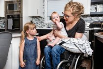 Una madre parapléjica en silla de ruedas hablando con su hija y sosteniendo a su bebé en su regazo mientras trabajaba en su cocina; Edmonton, Alberta, Canadá - foto de stock