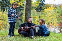 Молодой человек с синдромом Дауна играет на бубне, в то время как его отец играет на гитаре, а его мать поет вместе, наслаждаясь компанией друг друга в городском парке теплым осенним вечером: Эдмонтон, Альберта, Канада — стоковое фото