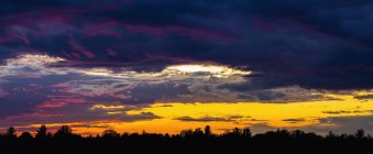 Dramatic sky at sunset with dark clouds and colourful light glowing above silhouetted trees; Bromont, Quebec, Canada — Stockfoto