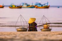 Una mujer sentada en la playa con vistas a los numerosos barcos de pesca en el agua frente a la costa, Ke Ga Cape; Ke Ga Island, Vietnam - foto de stock