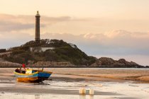 Colourful fishing boat tied to the beach and a lighthouse on a hill along the coast, Ke Ga Cape; Ke Ga, Vietnam — Stock Photo