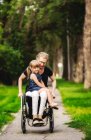 Una madre parapléjica sosteniendo a su niña en su regazo mientras la lleva a dar un paseo por la calle en su silla de ruedas en una cálida tarde de verano: Edmonton, Alberta, Canadá. - foto de stock
