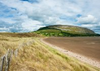 Irish coastline with beach grass and receded tide and old wooden fence, with a plateau mountain and cliffs in the background during summer; Strandhill, County Sligo, Ireland — Stock Photo