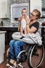 Une mère paraplégique tenant son bébé sur ses genoux, dans sa cuisine, alors qu'elle était assise dans son fauteuil roulant : Edmonton, Alberta, Canada — Photo de stock
