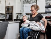 Une mère paraplégique tenant son bébé sur ses genoux, dans sa cuisine, tout en poussant dans son fauteuil roulant : Edmonton, Alberta, Canada — Photo de stock