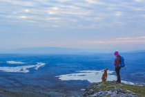 Lone woman hiker with knit hat and dog looking at each other on a cliff edge overlooking lakes in the distance on a cloudy evening in winter, Burren National Park; County Clare, Ireland — Stock Photo