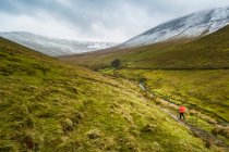 Самка-туристка в червоному плащі з червоним рюкзаком йде долиною, що веде до засніжених гір в похмурий зимовий день, гори Ґалті; графство Тіпперері, Ірландія. — стокове фото