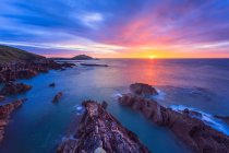 Sun rising over the Irish coastline with jagged rocks in the foreground and a lighthouse on an island in the distance; Ballycotton, County Cork, Ireland — Stock Photo