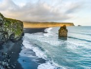 Cliffs and a rock formation along the coastline of the Southern Region of Iceland, with the surf washing up onto black sand in the foreground; Myrdalshreppur, Southern Region, Iceland — Stock Photo