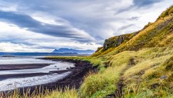Typische isländische Landschaft mit grüner Tundra, schwarzem Sand am Wasserrand und einer bergigen Region unter wolkenverhangenem Himmel; Island — Stockfoto