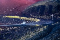Voiture voyageant sur une route sur un paysage volcanique noir avec une croix sur le bord de la route sur l'une des îles Westman ; Vestmannaeyjar, Région du Sud, Islande — Photo de stock