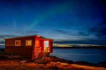 A small house on the water edge with a view of the tranquil coastline at nightfall and the glowing northern lights above; Nuuk, Sermersooq, Greenland — Stock Photo
