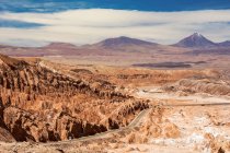 Дорога спускается в высокогорную пустынную долину с уникальными скальными образованиями слева и вулканической вершиной вдали; Сан-Педро-де-Атакама, Антофагаста, Чили — стоковое фото