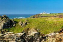 White lighthouse on top of hilly green fields framed by rocky outcrops and blue sky; Cornwall County, England — Stock Photo