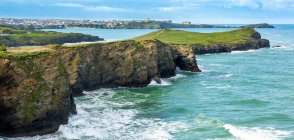 Penínsulas rochosas da costa do penhasco com campos verdes, céu azul e cidade no fundo distante; Condado de Cornwall, Inglaterra — Fotografia de Stock