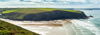 Rocky cliff shoreline peninsula with a patchwork of green fields, beaches and surf below, blue sky and clouds; Cornwall County, England — Stock Photo