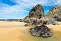 Playa arenosa con formaciones rocosas a lo largo de un acantilado rocoso con olas y cielo azul y nubes; Condado de Cornwall, Inglaterra - foto de stock