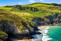 Falaises rocheuses le long du littoral avec collines herbeuses, ciel bleu et nuages ; comté de Cornwall, Angleterre — Photo de stock