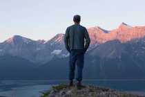 Hombre de pie en una cresta con vistas a un lago hacia los picos rocosos de la montaña; Kananaskis, Alberta, Canadá - foto de stock