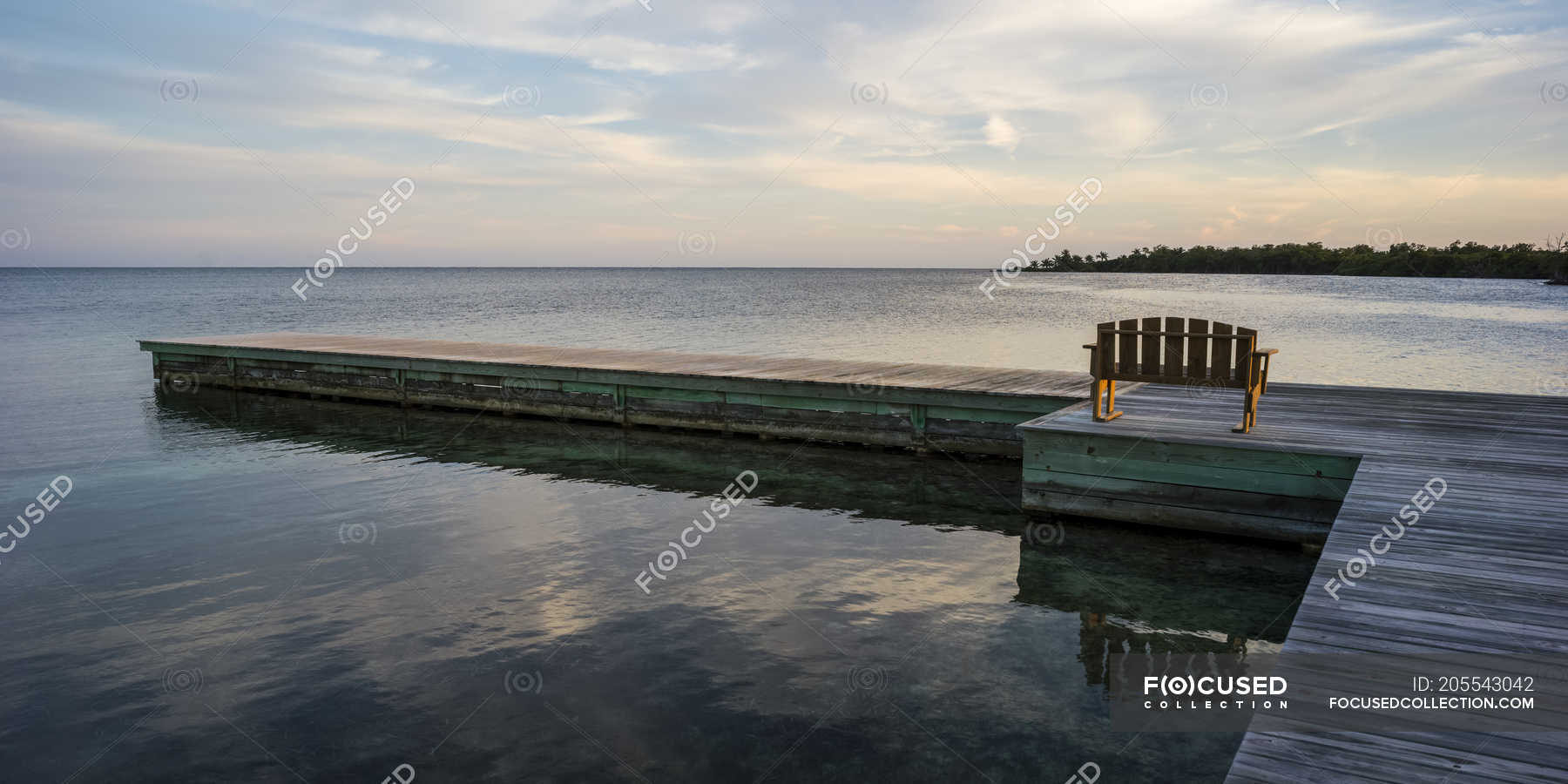 Wooden bench on a dock facing the coastline and open ocean at sunrise ...