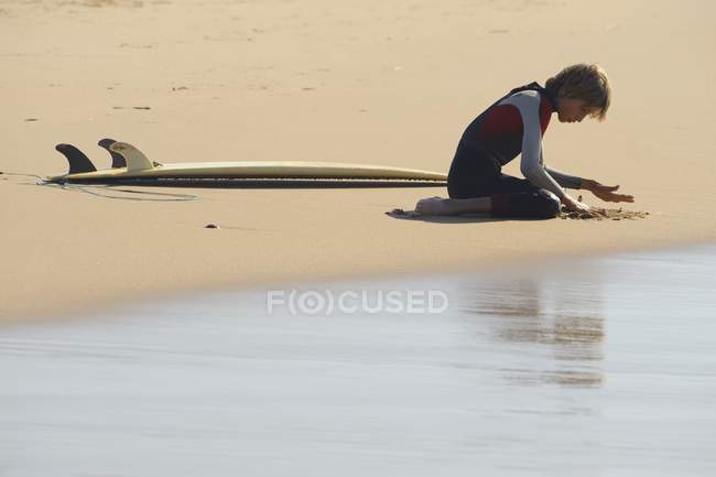Menino jogando a areia e sentado ao lado da prancha de surf — Fotografia de Stock