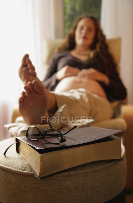 Mujer embarazada poniendo los pies arriba y y descansando con la biblia en hassock - foto de stock