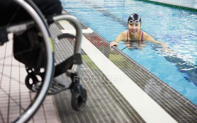 Mujer parapléjica nadando en piscina con silla de ruedas en el borde del agua - foto de stock