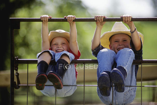 Menina jovem fazenda e menino vestindo chapéus de cowboy e botas de borracha balançar no portão de metal, pendurado por mãos e pernas — Fotografia de Stock