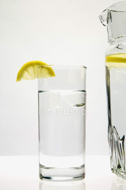 Стекло и кувшин воды с лимоном на белом — стоковое фото
