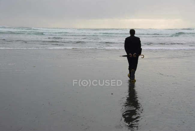Вид сзади человека на мокром пляже с зонтиком — стоковое фото