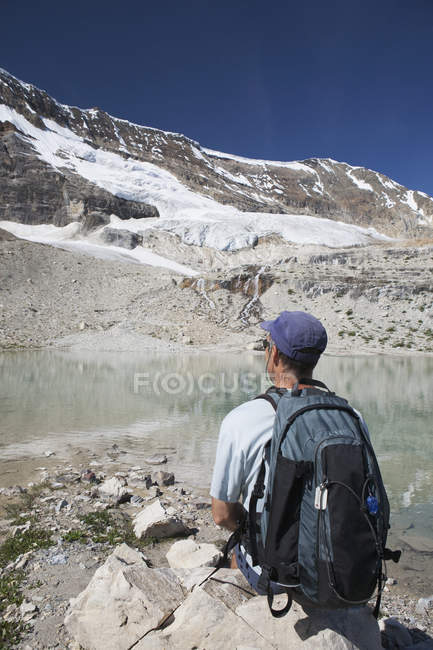 Caminhante Masculino sentado em uma rocha com geleira no lado da montanha fluindo para baixo em uma lagoa de montanha reflexiva com céu azul; Campo, Colúmbia Britânica, Canadá — Fotografia de Stock