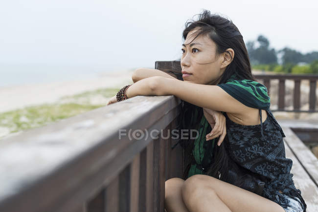 Jovem mulher sentada contra trilhos de madeira e olhando para o oceano — Fotografia de Stock