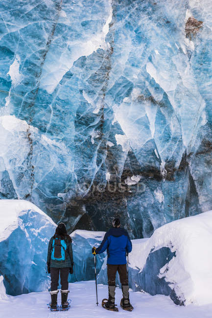 Couple of snowshoers examine large blue wall of ice with several collapsed pieces of ice at the bottom at the terminus of Canwell Glacier. Alaska, USA — Stock Photo