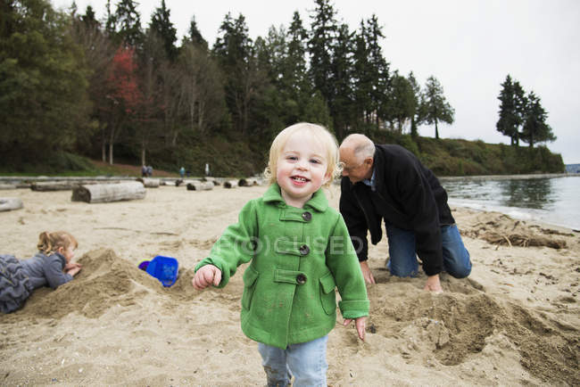 Una joven camina hacia la cámara mientras un abuelo y su nieta juegan en el fondo en la playa en invierno en Stanley Park; Vancouver, Columbia Británica, Canadá - foto de stock