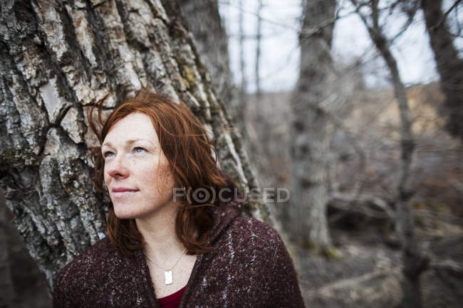 Retrato de una mujer con el pelo rojo contra un árbol y mirando contemplativo - foto de stock