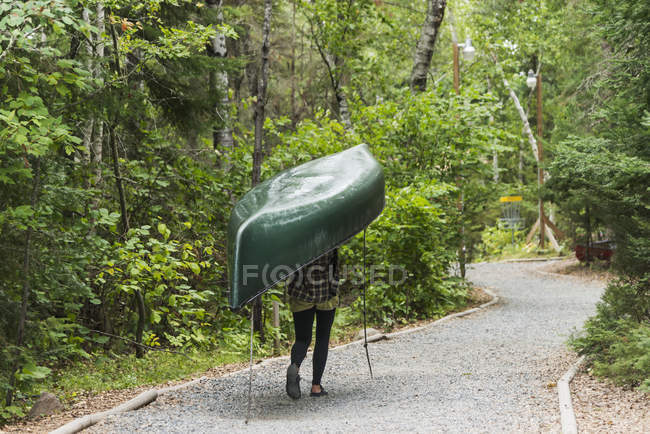 Una joven que lleva una canoa verde en su cabeza por un sendero; Ontario, Canadá - foto de stock