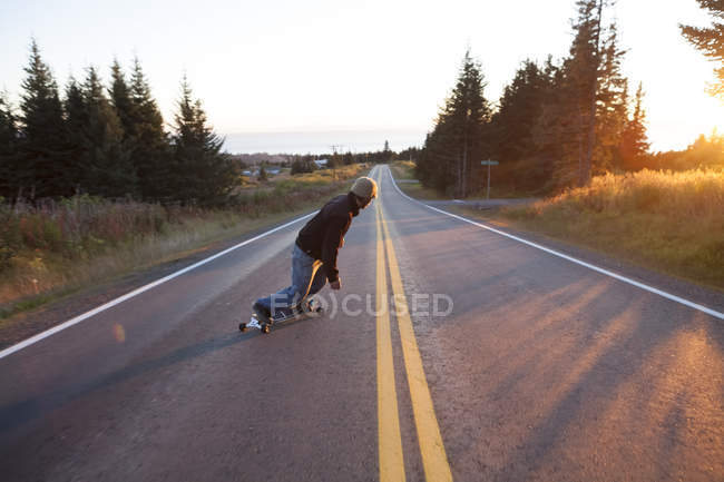 Вид сзади на молодого человека, катающегося на скейтборде по дороге в сумерках — стоковое фото